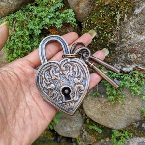 Puede incluir: Un candado de metal en forma de corazón con acabado en bronce antiguo y patrones de volutas en relieve en la parte frontal. El candado cuenta con una cerradura central y está unido a dos llaves de metal a juego en una pequeña argolla.