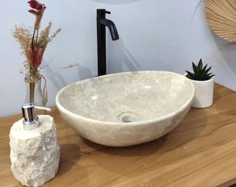 Puede incluir: Un lavabo ovalado beige con un grifo negro, un dispensador de jabón de piedra y una pequeña planta en maceta. El lavabo está sobre un mueble de madera con una pared azul claro al fondo. Flores secas decorativas en un jarrón de cristal.