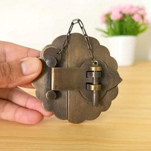Puede incluir: Un pestillo de armario circular de latón con borde festoneado y acabado cepillado. El herraje presenta un mecanismo de barra horizontal central con un pasador vertical, asegurado por una pequeña cadena de metal unida a la placa decorativa.