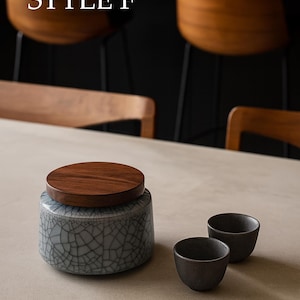 以下が含まれることがあります： 木製の蓋が付いた陶器の瓶と、2つの小さな濃い色のカップ。瓶はグレーのひび割れ釉薬です。「STYLE F」の文字が上部にあります。背景には木製の椅子が含まれています。