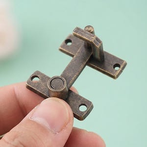 Könnte beinhalten: Ein kleiner Riegel aus Messing im antiken Stil mit einer verwitterten, dunklen Metalloberfläche. Das Beschlagteil besteht aus zwei rechteckigen Montageplatten mit jeweils zwei runden Schraublöchern, die durch einen Scharnierarm verbunden sind.