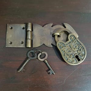 May include: A brass padlock with an ornate floral relief pattern, a matching hinged hasp plate featuring four screw holes, and two skeleton keys attached to a small metal ring. The metal components have a dark, aged patina finish.