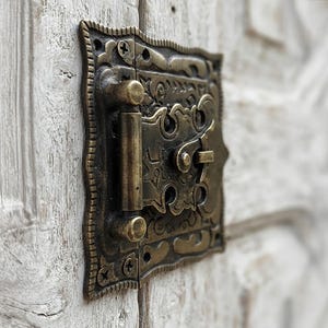 May include: An antique-style brass door latch with a rectangular backplate featuring intricate embossed patterns and a scalloped edge. The hardware includes a vertical cylindrical hinge and a central pivoting locking mechanism secured by screws.