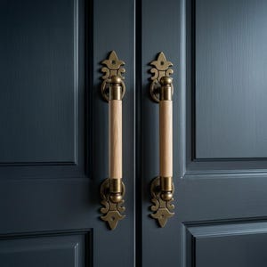 May include: Two ornate cabinet pulls with a vintage aesthetic. Each pull features a light wood handle, brass-colored hardware, and decorative flourishes. The pulls are mounted on a dark blue cabinet door, creating a classic look.