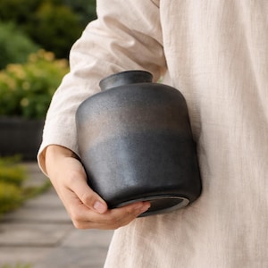 Könnte beinhalten: Eine dunkelgraue Keramikvase mit rundem Körper und schmalem Hals. Die Vase wird vor einer Person gehalten, die ein beiges Leinenhemd trägt. Die Vase hat eine matte Oberfläche und ein schlichtes, modernes Design.