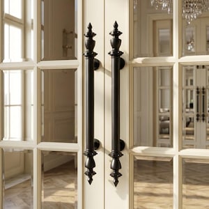 May include: Two black, ornate door handles with decorative finials are mounted on a cream-colored door. The door features multiple glass panes, reflecting the interior of a room with a chandelier.