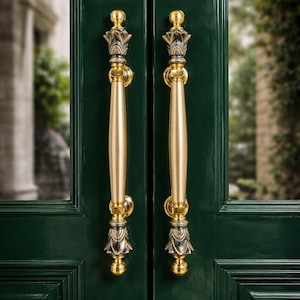 May include: Close-up of two gold-colored door handles with ornate, decorative detailing. The handles are mounted on a dark green door. The door handles have a long, cylindrical grip and are topped with a decorative crown-like design.
