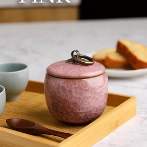 May include: A pink ceramic jar with a textured surface and a lid, featuring a bronze-colored handle. The jar sits on a bamboo tray with a wooden spoon and a light blue cup. The word "PINK" is displayed above the jar.
