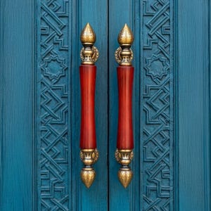 May include: Close-up of a teal blue door with two ornate, red and gold door handles. The handles have a teardrop-shaped finial and are set against a background of carved geometric patterns.