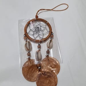 May include: A brown dreamcatcher with a woven web design, shell accents, and three large, round, brown shell pendants. The dreamcatcher is suspended by a brown string.