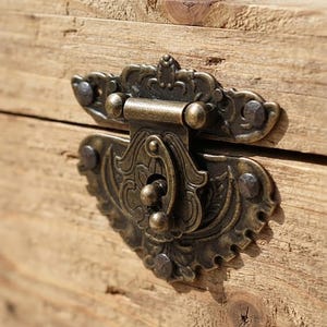 May include: A decorative antique-style brass latch with intricate scrollwork and embossed details, mounted onto a light brown natural wood surface. The metal hardware features a hinged clasp mechanism secured with small dark metal nails.