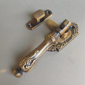 May include: Ornate antique bronze window handle and latch. The handle features a detailed, scrolling design and a curved lever. The latch is rectangular with two screw holes. The hardware is likely for a window or cabinet.