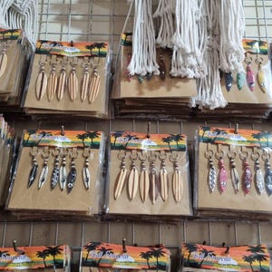 Puede incluir: Filas de llaveros empaquetados con diseños de tablas de surf en varios colores y patrones. Algunos llaveros son metálicos, mientras que otros tienen diseños pintados. Cuerda blanca cuelga en el fondo, y el embalaje tiene un tema tropical.