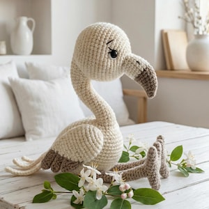 May include: A crocheted flamingo toy in cream and brown, with a long neck and curved beak. The handmade bird is sitting on a white wooden table, decorated with white flowers and green leaves. The toy is approximately 12 inches tall.
