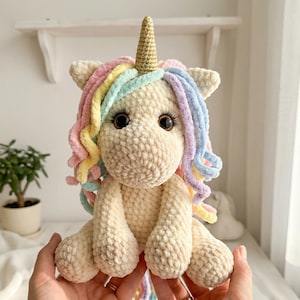 May include: A cream-colored, crocheted unicorn plush toy with a golden horn and rainbow-colored mane. The unicorn has large, dark eyes and is sitting upright. The plush is soft and cuddly, perfect for a child's bedroom or nursery.