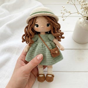 Könnte beinhalten: Eine handgefertigte Häkelpuppe mit braunen Locken, die ein salbeigrünes Kleid, eine kleine braune Handtasche und einen cremefarbenen Hut trägt. Die Puppe hat braune Schuhe und weiße Socken. Die Puppe wird gehalten.