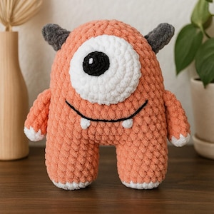 May include: A handmade, orange crochet monster with one large eye, two small horns, and a smiling mouth. The monster has white accents on the eye, feet, and hands. This plush toy is a cute and cuddly handmade item.
