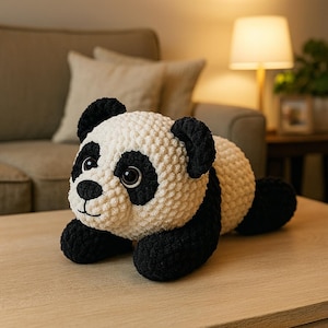May include: A plush panda bear toy with a textured, cream-colored body and black accents on the ears, eyes, and limbs. The panda has large, round eyes and is lying on a light-colored wooden surface.