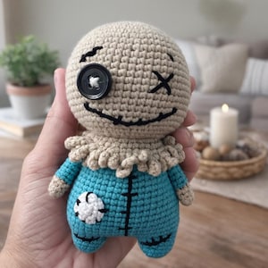 May include: A handmade crochet doll with a tan head, black button eye, and stitched mouth and cross-shaped eye. The doll wears a blue outfit with a white flower patch. The doll is held in a hand.
