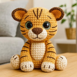 Peut inclure: Une peluche tigre au crochet, faite à la main. Le tigre est de couleur jaune et marron, avec des rayures noires. Il a de grands yeux verts et noirs scintillants, un nez marron et de petites oreilles. Il est posé sur une surface en bois.