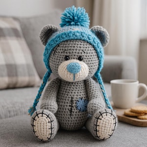 May include: A handmade gray crochet teddy bear wearing a blue hat with a pom-pom and braided ties. The bear has a white muzzle and paws, a blue nose, and a blue patch. The bear is sitting on a gray surface.