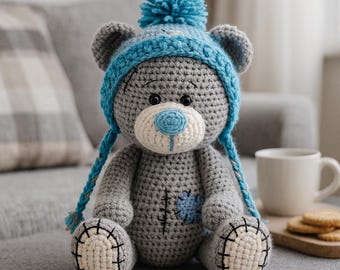 Peut inclure: Un ours en peluche crocheté à la main, de couleur grise, portant un bonnet bleu avec un pompon et des tresses. L'ours a un museau et des pattes blancs, un nez bleu et une pièce bleue. L'ours est assis sur une surface grise.