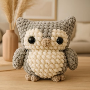May include: A handmade, gray, white, and beige crochet owl. The owl has large black eyes and a small beige beak. The body is a mix of white and beige, with gray wings and head. The owl is sitting on a wooden surface.