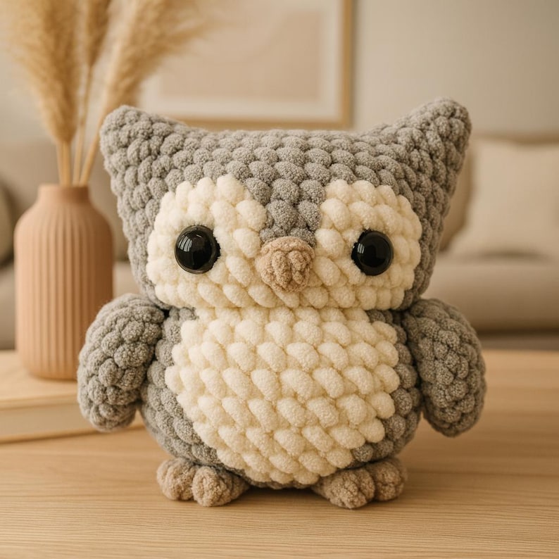 May include: A handmade, gray, white, and beige crochet owl. The owl has large black eyes and a small beige beak. The body is a mix of white and beige, with gray wings and head. The owl is sitting on a wooden surface.
