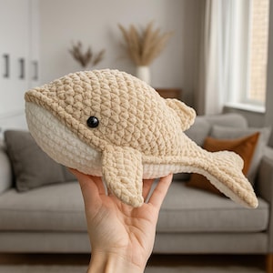 May include: A beige crocheted whale plush toy with a white belly and black eyes. The whale has fins and a tail, and is held in a hand. The plush toy is made of a soft, textured yarn.