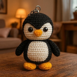 May include: A crocheted penguin keyring. The penguin is black and white with yellow feet and an orange beak. It has large black eyes and is attached to a silver metal keyring. The penguin is approximately 10 cm tall.