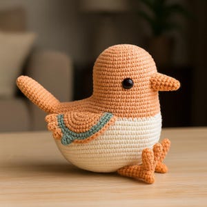 May include: A handmade crochet bird in shades of orange and cream. The bird has a rounded body, a small beak, and a dark eye. The wings and feet are also orange, and there is a green and blue accent on the bird's back. The bird is sitting on a wooden surface.