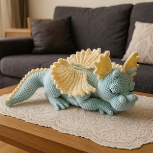 Könnte beinhalten: Ein hellblaues gehäkeltes Dinosaurier-Plüschtier mit gelben Akzenten, einschließlich Flügeln und Kopfteilen. Das handgefertigte Stofftier liegt auf einem Tisch mit einer Spitzendecke. Der Dinosaurier hat geschlossene Augen.