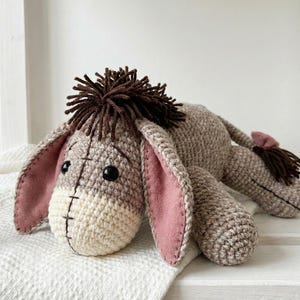 May include: A handmade crochet donkey plush toy in shades of beige, brown, and pink. The donkey has large, floppy ears with pink felt lining, a brown mane, and black button eyes. The toy is lying down on a white surface.
