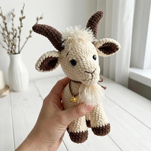 May include: A crocheted goat figurine with a cream-colored body, brown horns, and dark brown hooves. The goat has a small gold bell around its neck. The handmade toy is held in a person's hand.