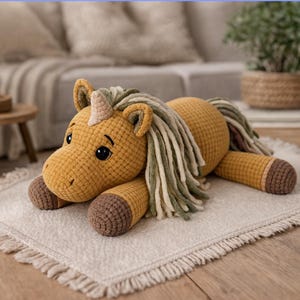 May include: A handmade crochet unicorn plush toy in shades of yellow, brown, and cream. The unicorn has a horn, long mane, and tail. It is lying on a white rug with a fringed edge. The toy is soft and cuddly.