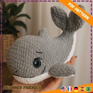 May include: A crocheted gray whale plushie with white accents on the belly and mouth. The whale has a smiling face with black eyes and a small black line for a smile. The image includes the text "PDF PATTERN" and "BEGINNER FRIENDLY".