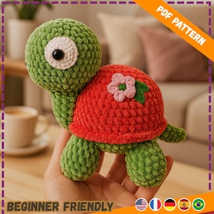 Può includere: Una tartaruga all'uncinetto verde e rossa fatta a mano con un fiore sul guscio. La tartaruga ha un grande occhio bianco e nero. L'immagine include il testo "PDF PATTERN" e "BEGINNER FRIENDLY".