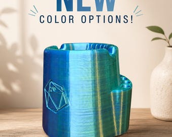 May include: A blue and green ashtray with a geometric design and the text "NEW COLOR OPTIONS!". The ashtray is made of a 3D-printed material and sits on a wooden surface.