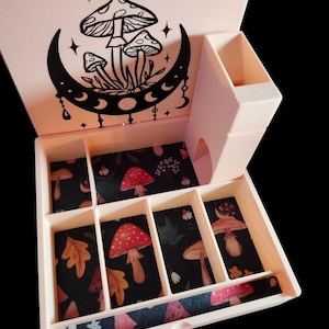 Puede incluir: Una caja de joyería rosa claro con un diseño de hongo y luna creciente negro en la tapa. El interior presenta compartimentos con un forro de tela negra adornado con patrones de hongos y hojas. Un pequeño compartimento adjunto está en el lateral.