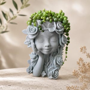 May include: A gray concrete planter shaped like a human head with closed eyes and a hand resting on the cheek. The head is adorned with sculpted roses and leaves, and it contains a trailing string of pearls plant with small green spheres.