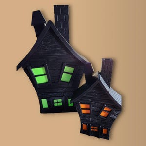 May include: Two black, textured, haunted house decorations with glowing green and orange windows. The houses have chimneys and are set against a tan background. The larger house has green windows, while the smaller house has orange windows.