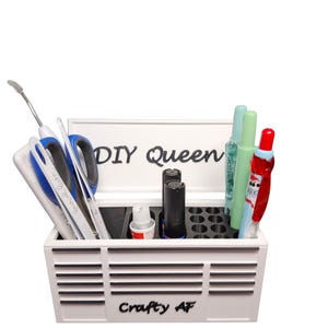 May include: White craft organizer box with the text "DIY Queen" and "Crafty AF". The box contains various craft supplies, including scissors, pens, and bottles. The box has a slotted design on the front.