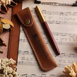 May include: A brown leather pen case with a gold button clasp, personalized with the name "Nathaniel" and the date "October 16, 2025". A burgundy and gold pen rests beside the case on sheet music. Decorative elements surround the pen and case.