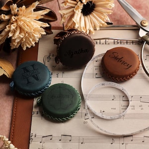 May include: Several personalised leather tape measures in shades of brown, green, and teal. Each has a name engraved, including "Agatha," "Sidney," "Nadine," and "Charlotte." The image also shows scissors, sheet music, and dried flowers.