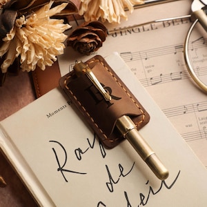 Op de afbeelding: Een messing pen met een leren etui, rustend op een open boek met de woorden "Ray de Noir". De pen ligt naast bladmuziek en gedroogde bloemen, wat een vintage esthetiek creëert.