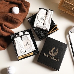 May include: Two black gift boxes containing white golf gloves with black trim and wooden tees. One box features the Maynard logo, while the other shows the Teofilo logo. A golf club, two white golf balls, and a brown knit fabric are also present.