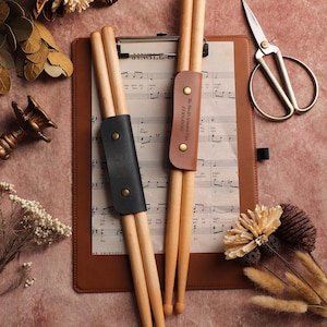 Puede incluir: Dos pares de baquetas de madera con fundas de cuero, una negra y otra marrón, descansan sobre un portapapeles con partituras. La funda marrón tiene grabado el texto "The World's Greatest Dad". Un par de tijeras doradas y flores secas completan el conjunto.