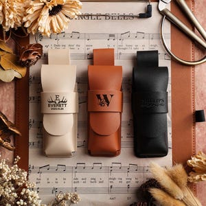 Puede incluir: Tres fundas de cuero en crema, marrón y negro, cada una con una inscripción personalizada. La funda crema dice "EVERETT 2025.10.24". La funda marrón tiene la inicial "W", y la funda negra dice "PHILLIPS". Las fundas están sobre partituras.