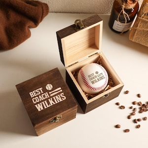 May include: A dark brown wooden box with a metal latch, featuring the engraved text "BEST COACH WILKINS" and a baseball icon. Inside the open box sits a white baseball with red stitching, also printed with "BEST COACH WILKINS" and a baseball icon.