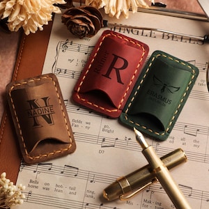 May include: Three leather pen holders in brown, red, and green, each with a unique design and text. The brown holder has the name "NADINE" and the letter "N". The red holder has the letter "R" and the name "Rainer". The green holder has the name "ERASMUS".